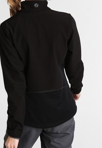 Chaqueta negra con un cuello alto, hecha de una tela suave y texturizada; presenta costuras contrastantes y un logotipo en el cuello trasero.