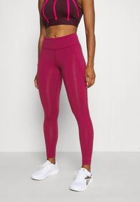 Träningsbehå i vinröd färg med rosa detaljer, kombinerad med högmidjade, stretchiga magenta leggings. Modellen bär vita träningsskor.