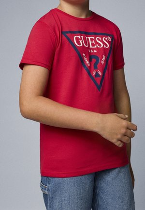 Enfant portant un t-shirt rouge à manches courtes avec un grand logo Guess et un jean bleu délavé sur fond gris.