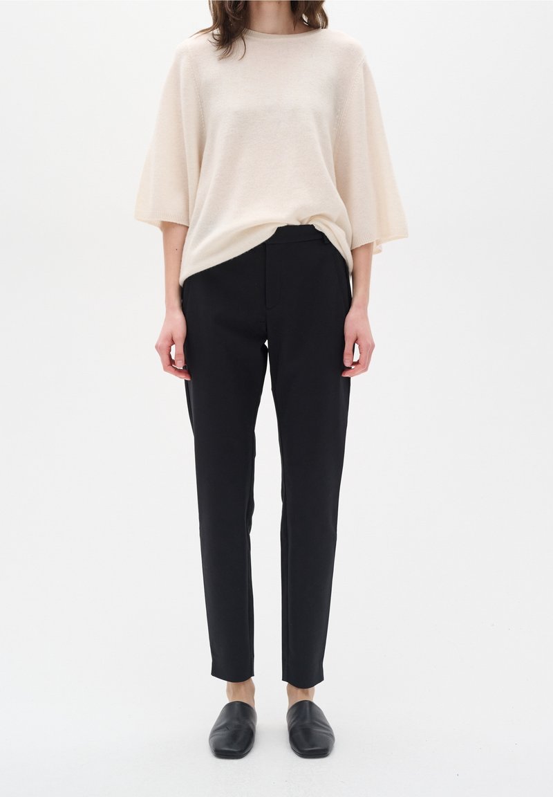 Femme portant un haut ample couleur crème, un pantalon noir ajusté longueur cheville et des chaussures noires sans lacets, debout devant un fond blanc.