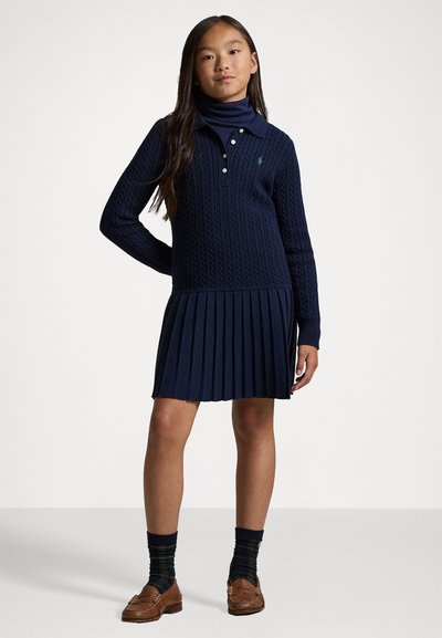 Ναυτικό φόρεμα με πλέξη καλωδίου, ψηλό λαιμό και πλισέ φούστα. Εμπρός με τρία κουμπιά και ραμμένο μανίκι. Συνδυάζεται με καφέ loafers και καλλιτεχνικές κάλτσες.