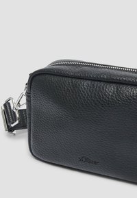 Zwarte leren tas met een gestructureerde oppervlakte, ritssluiting en metalen hardware. Voorzien van een hengsel om te dragen, gelabeld met "s.Oliver."