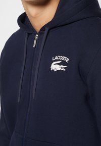 Sweat à capuche zippé bleu marine avec cordons, présentant un logo Lacoste blanc brodé avec un crocodile sur la poitrine gauche.