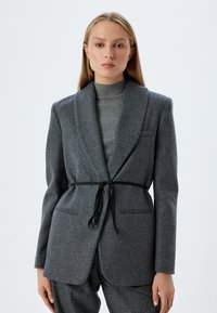 Blazer en laine gris avec un col châle, doté d'une ceinture noire à la taille et de poches avant. Le modèle porte un pull à col roulé gris côtelé en dessous.