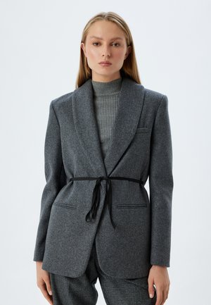 Blazer en laine gris avec un col châle, doté d'une ceinture noire à la taille et de poches avant. Le modèle porte un pull à col roulé gris côtelé en dessous.