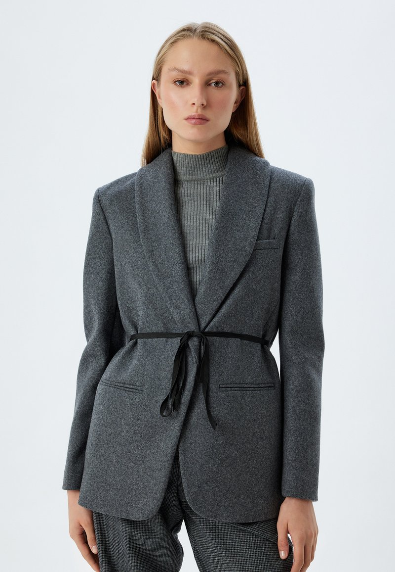 Grå uldblazer med sjalskrave, der har et sort slipsbælte i taljen og frontlommer. Modellen har en ribbet grå turtleneck på under.