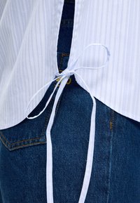 Λευκό ριγέ πουκάμισο με λεπτομέρεια γραβάτας στο πίσω μέρος συνδυασμένο με μπλε τζιν, που δείχνει ραφές και λεπτομέρειες τσέπης.