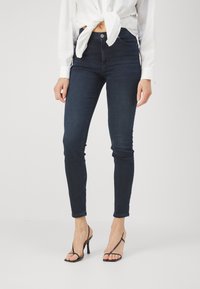 Jeans ajustados de color azul oscuro con una cintura alta, un desvanecimiento sutil y un diseño clásico de cinco bolsillos, combinados con tacones negros de tiras.