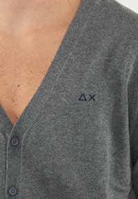 Cardigan grigio con scollatura a V profonda, caratterizzato da bottoni viola e un piccolo logo ricamato. Texture morbida, tessuto lavorato a maglia con una vestibilità classica.