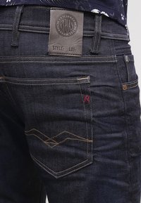 Mörkblå denimjeans med en läderpatch bak, gula sömnadsdetaljer och en dekorativ design på bakfickan.