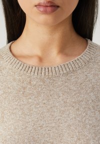 Pull beige en maille avec un col rond côtelé et une texture douce. Le tissu présente un motif subtil de petites taches sur toute la surface.