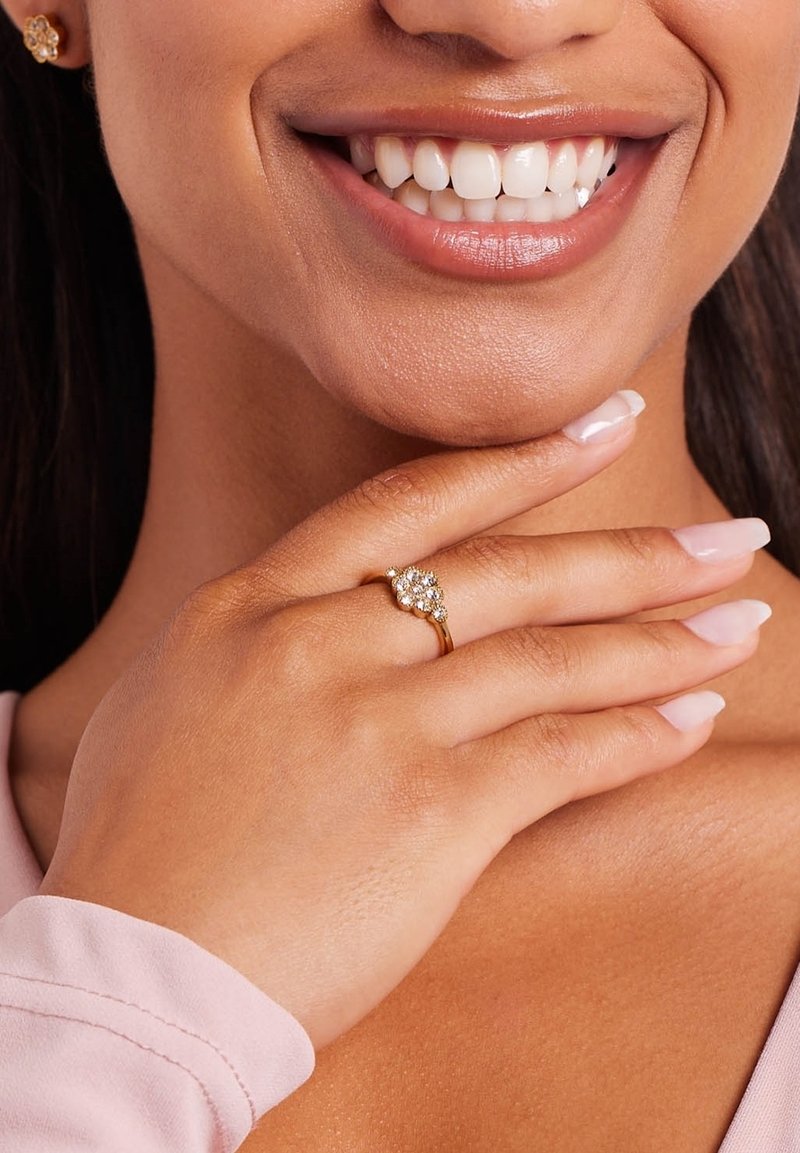 Gouden ring met een cluster van heldere stenen, vastgehouden door een hand met lange, netjes manicured nagels, tegen een zachte roze stoffen achtergrond.