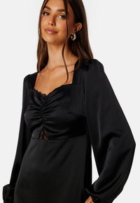 Černé saténové šaty se čtvercovým výstřihem, s nařaseným předním dílem, krajkovými detaily a dlouhými nadýchanými rukávy. Hladká textura a elegantní silueta.