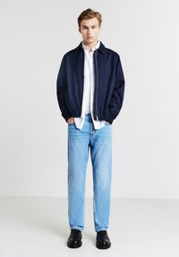 Giubbotto bomber blu navy con zip, camicia bianca sotto, jeans azzurri chiari e scarpe in pelle nere. Design semplice e vestibilità rilassata.