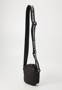 Svart crossover-bag laget av glatt materiale, med rektangulær form, glidelås og justerbar, merket stropp.