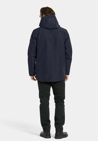 Marineblauwe waterdichte jas met capuchon, gestructureerde stof, elastische manchetten en een rechte snit. Gecombineerd met zwarte broek en schoenen met rubberen zolen.