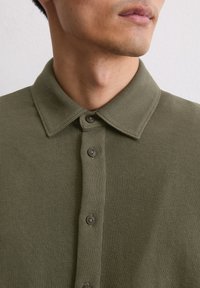Polo shirt verde oliva, a costine, con colletto a bottoni e cinque bottoni scuri sulla patta. Tessuto testurizzato con una vestibilità aderente.