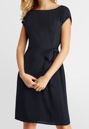Femme portant une robe noire longueur genou avec des manches courtes et un nœud à la taille sur le côté, debout, mains jointes devant.