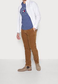 Bílá košile s knoflíky, modré tričko s potiskem, hnědé kordové kalhoty a béžové kotníkové boty. Klíčové prvky: barva, materiál a střih.