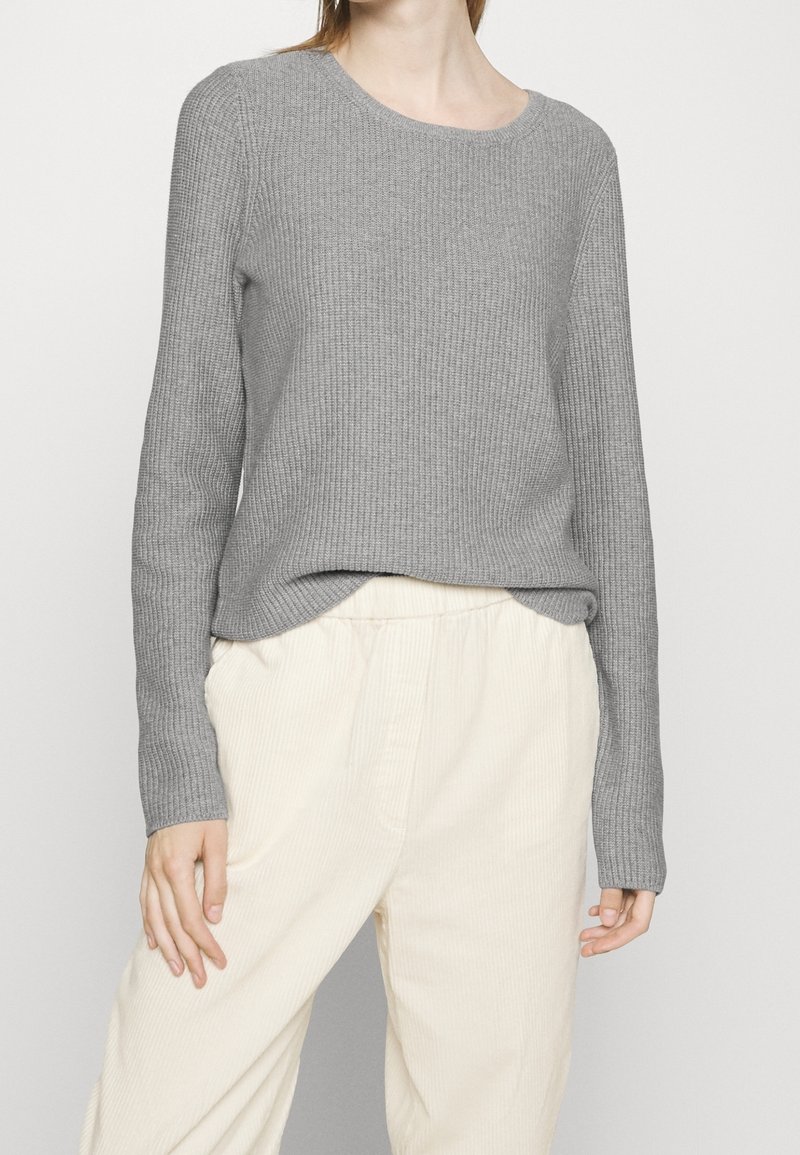Personne portant un pull gris côtelé à manches longues, légèrement rentré dans un pantalon ample taille haute beige clair sur un fond uni.