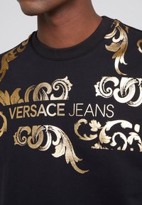Sweatshirt noir avec des motifs floraux dorés et le texte "VERSACE JEANS", col rond, texture douce et motifs détaillés.