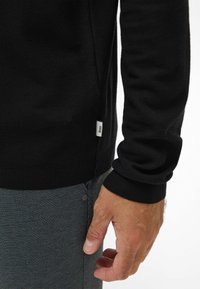 Maglione lavorato a maglia nero con polsini a coste, dotato di un'etichetta laterale. La mano è appoggiata su un tessuto grigio testurizzato dei pantaloni, mostrando i dettagli e la vestibilità.