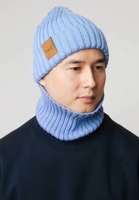 Jasnoniebieska dzianinowa czapka z prążkowaną strukturą i brązową skórzaną naszywką, zestawiona z dopasowanym prążkowanym ocieplaczem na szyję w tym samym kolorze.