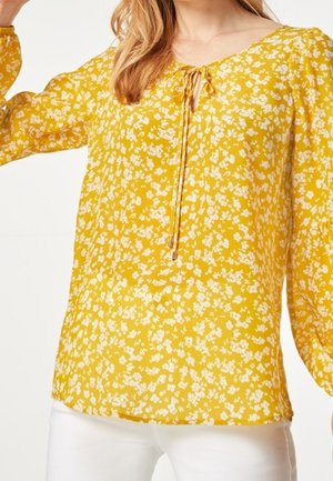 Blusa amarilla con estampado floral, de manga larga, tela ligera, cuello con lazo y patrón de flores blancas dispersas. Combinada con pantalones blancos.