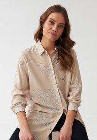 Beige Bluse mit langen Ärmeln aus leichtem Material, mit einem subtilen grauen Blumenmuster. Knöpfe an der Vorderseite und klassischer Kragen.