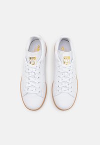 Zapatillas de cuero blanco con suela de goma, que presentan un diseño perforado, cordones planos y un logo dorado en la lengüeta.