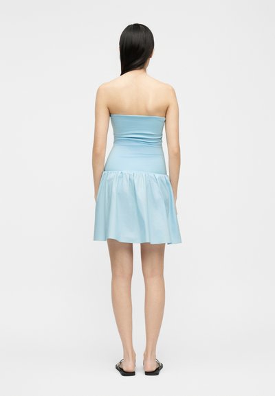 Γυναίκα με μακριά σκούρα μαλλιά που φοράει αμάνικο ανοιχτό γαλάζιο φόρεμα και μαύρα επίπεδα σανδάλια, στέκεται πλάτη σε ένα λευκό φόντο.