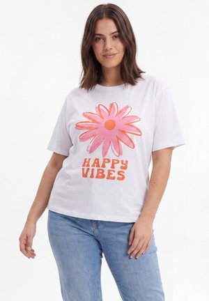 Vrouw met bruin haar die een wit t-shirt draagt met een grote roze en oranje bloem en de tekst "HAPPY VIBES", gecombineerd met lichtblauwe jeans.