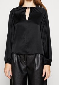 Donna che indossa una blusa nera di raso a maniche lunghe con scollatura a chiave e pantaloni in pelle neri a vita alta.