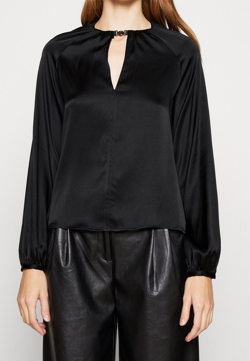 Donna che indossa una blusa nera di raso a maniche lunghe con scollatura a chiave e pantaloni in pelle neri a vita alta.