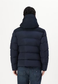 Temno modra puffer jakna z visokim ovratnikom, ki ima prešit dizajn in gladko zunanjost. Toploto poudarja debela, mehka podloga.