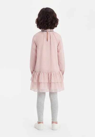 Jeune enfant aux cheveux bouclés, de dos, portant une robe rose à volants, un legging gris et des chaussures blanches sur un fond uni.