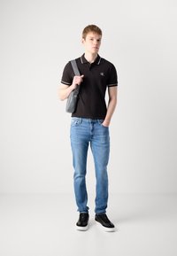 Jauns vīrietis, kurš valkā melnu polo kreklu, gaiši zilos džinsos un melnās kedās, vienā plecā nes pelēku plecu somu uz baltā fona.