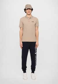 Beige polo skjorte med korte ermer og krage, kombinert med marineblå joggebukser med hvit logo-print, og en matchende caps. Hvite sneakers.
