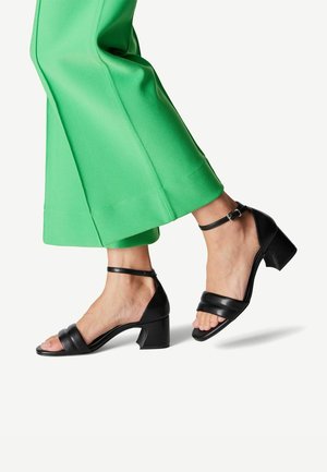 Sandales à talons hauts en cuir noir avec une sangle autour de la cheville et deux larges bandes pour les orteils ; exposées à côté d'un pantalon vert à jambes larges.