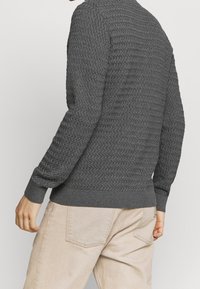 Grå stickad tröja med ett strukturerat mönster, rund halsringning och ribbade muddarna och fållen, tillsammans med ljusbeiga byxor.