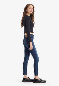 Mujer joven con cabello rizado largo que lleva una camiseta de manga larga negra, jeans ajustados azul oscuro y mocasines negros, de pie de perfil.