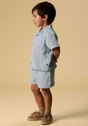 Băiat tânăr îmbrăcat într-o cămașă cu mânecă scurtă texturată, de culoare albastru deschis, și pantaloni scurți, cu șosete bej și pantofi maro în stil mocassin, stând lateral cu mâinile la spate.