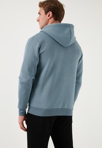 Sudadera azul claro hecha de una tela suave y texturizada. Presenta un bolsillo tipo canguro y una capucha con cordón, con puños y dobladillo acanalados.