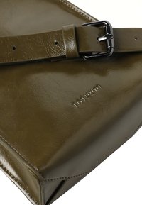 Olijfgroene leren tas met glanzende afwerking, voorzien van een gestructureerde vorm, een gespband en een gegraveerde merknaam aan de voorkant.