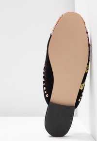 Semelle de chaussure plate en beige et noir avec des clous dorés et des détails en tissu floral, photographiée par le dessous sur fond blanc.