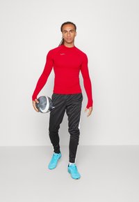 Rotes langärmliges Sportshirt mit taillierter Passform, kombiniert mit schwarzen Hosen mit weißen Seitenstreifen. Hält einen grauen Fußball und trägt blaue Schuhe.