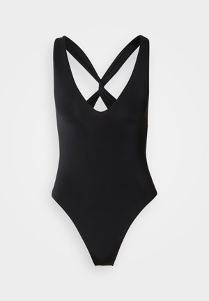 Juodas vientisas maudymosi kostiumėlis su gilia V formos iškirpte ir sukryžiuotomis nugaros petnešėlėmis ant šviesiai pilkos spalvos fono.