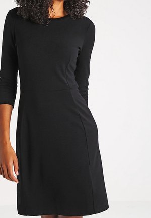 Woman wearing a simple black knee-length dress with three-quarter sleeves standing against a plain white background.