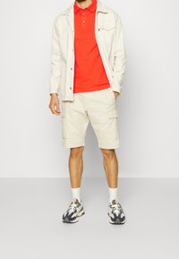 Beżowe szorty cargo, czerwona koszulka polo, lekka kurtka oraz wzorzyste trampki w niebieskie i kremowe wzory. Szorty mają boczne kieszenie i luźny krój.