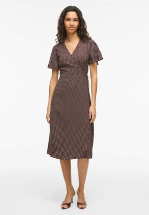 Femeie cu păr creț purtând o rochie maro închis, până la genunchi, cu buline albe și mâneci scurte evazate, stând desculță.